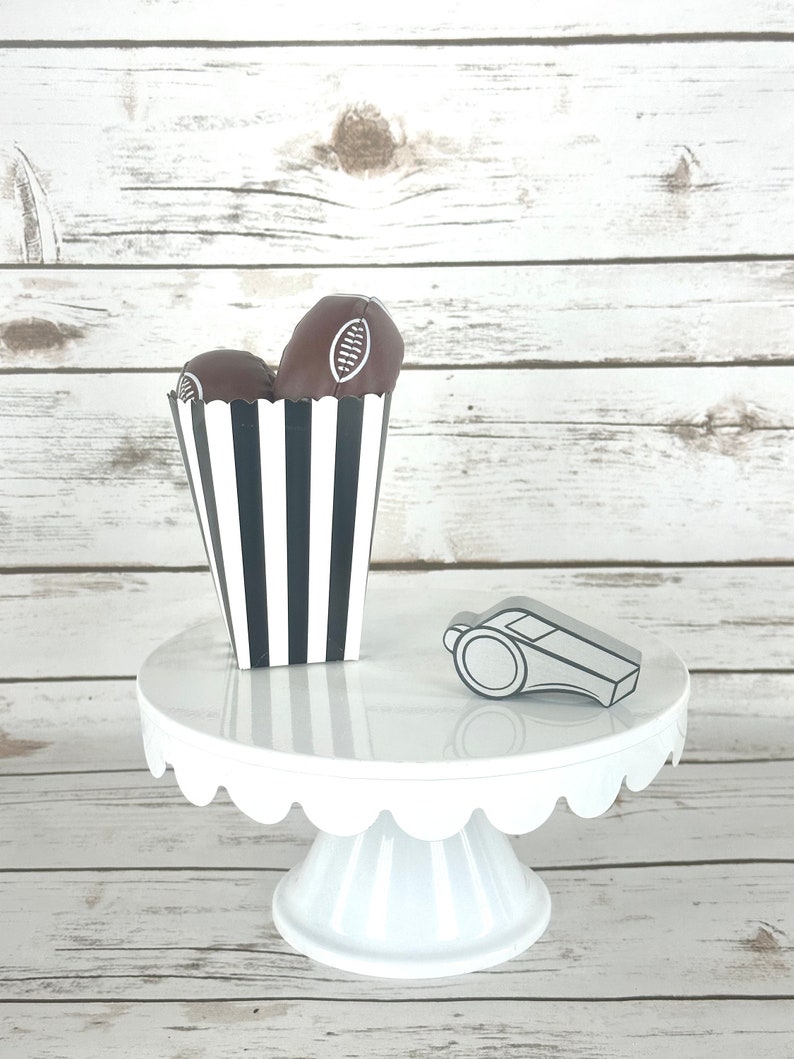 May include: A white cake stand with a scalloped edge, decorated with a black and white striped box filled with chocolate footballs and a silver whistle.