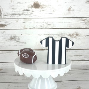 May include: A white cake stand with a scalloped edge, decorated with two brown footballs and a black and white striped referee's jersey.