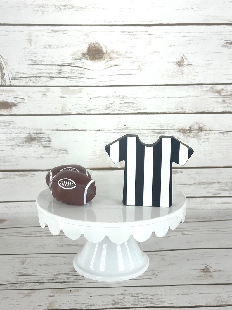 May include: A white cake stand with a scalloped edge, decorated with two brown footballs and a black and white striped referee's jersey.