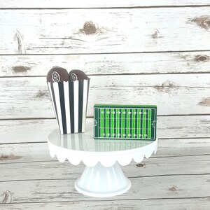 May include: A white cake stand with a scalloped edge, decorated with two brown footballs in a black and white striped paper cup and a green and white football field.