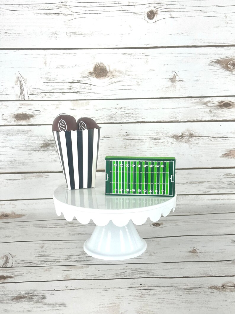 May include: A white cake stand with a scalloped edge, decorated with two brown footballs in a black and white striped paper cup and a green and white football field.