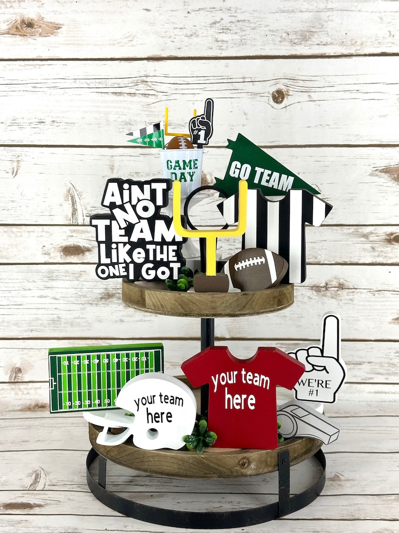 May include: A two-tiered wooden tray with a black metal stand. The top tier features a football, a football goal post, a referee's jersey, a pennant that reads "Go Team", and a sign that says "Ain't No Team Like The One I Got". The bottom tier has a football helmet, a football field, a red jersey that says "Your Team Here", a whistle, and a foam finger that says "We're #1".