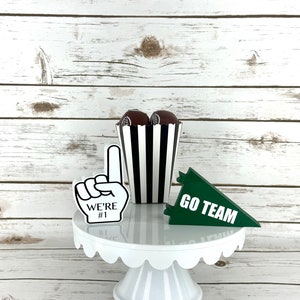 May include: White cake stand with a scalloped edge, decorated with a black and white striped popcorn container, a green pennant that says "GO TEAM", and a foam finger that says "WE'RE #1".