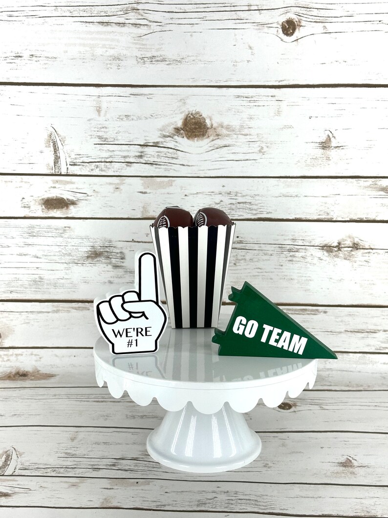 May include: White cake stand with a scalloped edge, decorated with a black and white striped popcorn container, a green pennant that says "GO TEAM", and a foam finger that says "WE'RE #1".