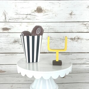 May include: A white cake stand with a scalloped edge, decorated with two chocolate footballs in a black and white striped paper cup and a yellow miniature football goal.