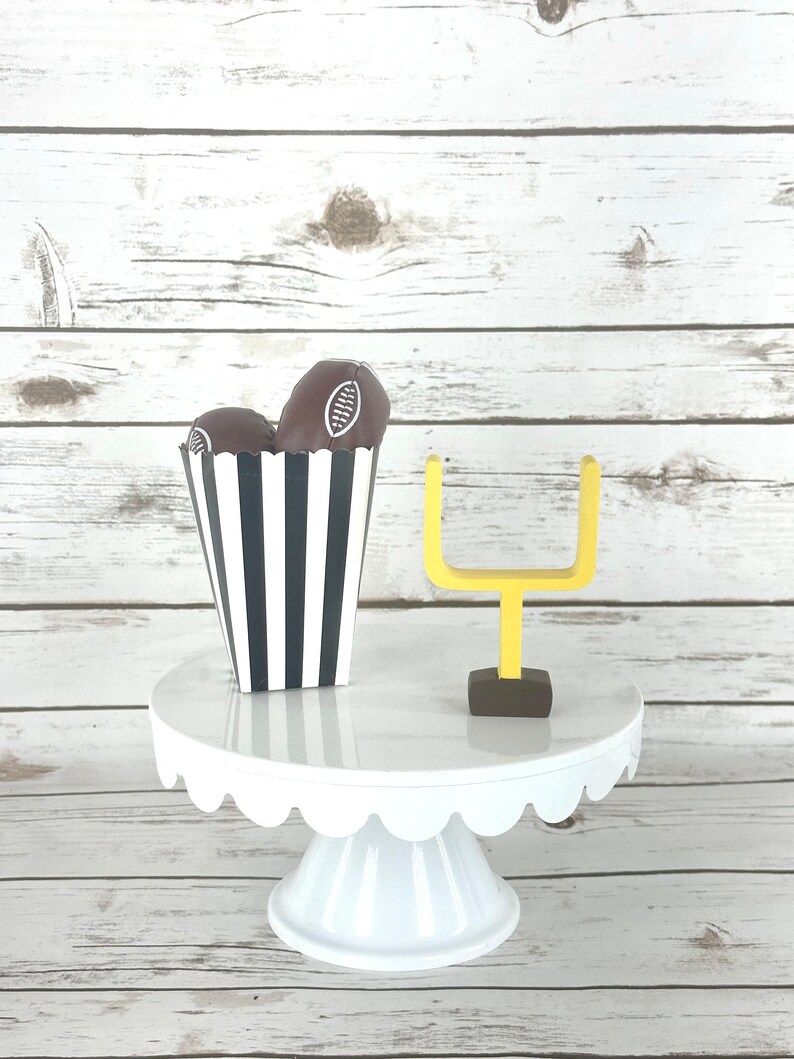 May include: A white cake stand with a scalloped edge, decorated with two chocolate footballs in a black and white striped paper cup and a yellow miniature football goal.