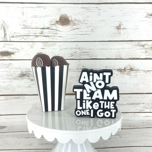 May include: A white cake stand with a black and white striped box filled with chocolate footballs. The box is next to a black sign that reads "Ain't no team like the one I got".