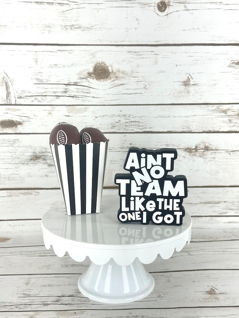 May include: A white cake stand with a black and white striped box filled with chocolate footballs. The box is next to a black sign that reads "Ain't no team like the one I got".