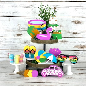 May include: A tiered tray decorated with summer-themed wooden cutouts. The top tier features a pink flamingo pool float, a yellow and blue pool ring, a green and orange popsicle, and a white swimsuit with green and orange flowers. The middle tier has a pair of yellow and orange flip flops, a yellow and blue beach ball, a purple and white umbrella, and a green and orange popsicle. The bottom tier has a pink and white car with flowers, a pink and white ice cream cone, two blue and white snow cones, and a pair of black sunglasses with palm trees.