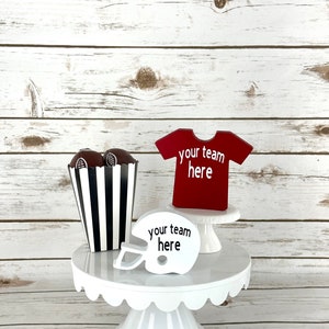 May include: White cake stand with a black and white striped popcorn container, two brown footballs, a red football jersey with the text "your team here", and a white football helmet with the text "your team here".
