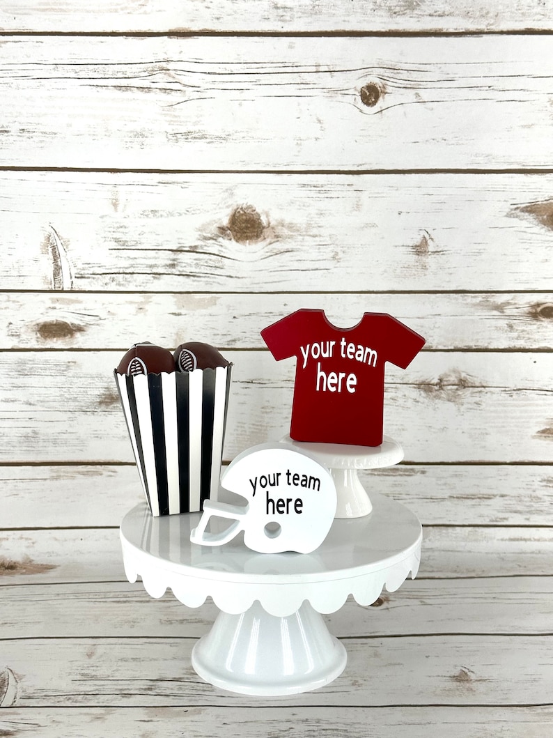 May include: White cake stand with a black and white striped popcorn container, two brown footballs, a red football jersey with the text "your team here", and a white football helmet with the text "your team here".