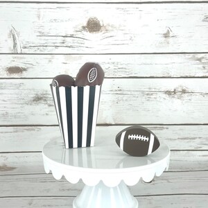 May include: A white cake stand with a scalloped edge holds a black and white striped box filled with chocolate football-shaped treats. A brown football with white laces sits on the stand.