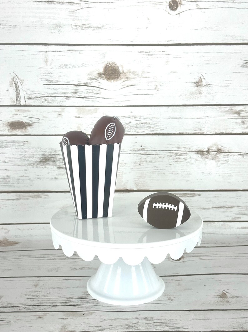 May include: A white cake stand with a scalloped edge holds a black and white striped box filled with chocolate football-shaped treats. A brown football with white laces sits on the stand.