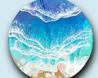 May include: A round resin art piece depicting a blue ocean scene with white foamy waves crashing on a sandy beach with seashells.