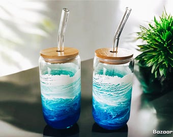 Könnte beinhalten: Zwei klare Glasbecher mit Bambusdeckeln und Glasstrohhalmen. Die Becher sind mit einem blauen und weißen Ozeanwellenmuster verziert.