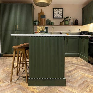 Puede incluir: Una isla de cocina con un exterior verde acanalado y una encimera blanca. Tres taburetes de bar de madera est&aacute;n colocados frente a la isla. La cocina cuenta con armarios verdes, una l&aacute;mpara colgante dorada y un suelo de madera en espiga.