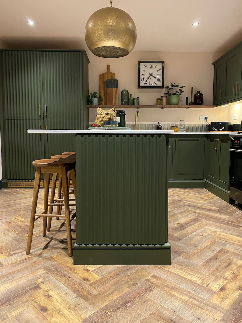 Puede incluir: Una isla de cocina con un exterior verde acanalado y una encimera blanca. Tres taburetes de bar de madera est&aacute;n colocados frente a la isla. La cocina cuenta con armarios verdes, una l&aacute;mpara colgante dorada y un suelo de madera en espiga.