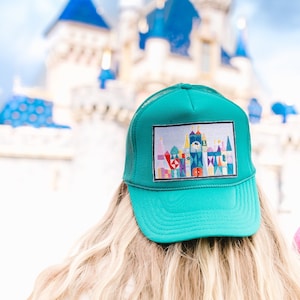 以下が含まれることがあります： 後ろが白いメッシュになっているターコイズブルーのトラッカーハット。帽子には、時計塔のある城がカラフルに刺繍されたパッチが付いています。