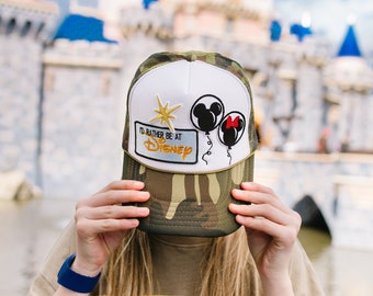 Może przedstawiać: Czapka trucker w kamuflażu z białym panelem z przodu. Czapka ma naszywkę z napisem "I'd rather be at Disney" z motywem balonu z postacią Myszki Miki i Minnie Mouse. Czapka jest trzymana przed twarzą osoby, z rozmytym obrazem zamku w tle.