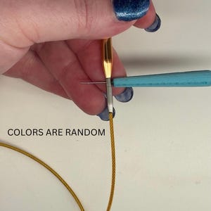 Könnte beinhalten: Eine goldfarbene Stricknadel mit einem flexiblen goldenen Kabel, das an einem silberfarbenen Metallverbinder befestigt ist. Ein hellblaues Werkzeug wird in den Verbinder eingeführt, um ihn festzuziehen. Der Text "COLORS ARE RANDOM" steht auf dem Bild.