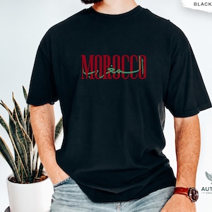 May include: A black short-sleeved t-shirt featuring the word MOROCCO printed in red capital letters across the chest, with Arabic script layered in green over the text. The shirt has a crew neckline and a standard fit, shown in a solid black color.