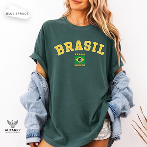 May include: A blue spruce colored t-shirt featuring the yellow text BRASIL in an arched font above a graphic of the Brazilian flag with five yellow stars and the text WORLD CUP 2026 printed in yellow underneath the flag design.