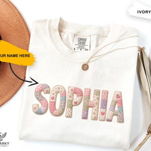 May include: An ivory short-sleeved t-shirt with the name SOPHIA appliqued across the chest in a patchwork fabric featuring various floral and gingham patterns in shades of pink, blue, and green. The shirt has a crew neckline and a Comfort Colors label.