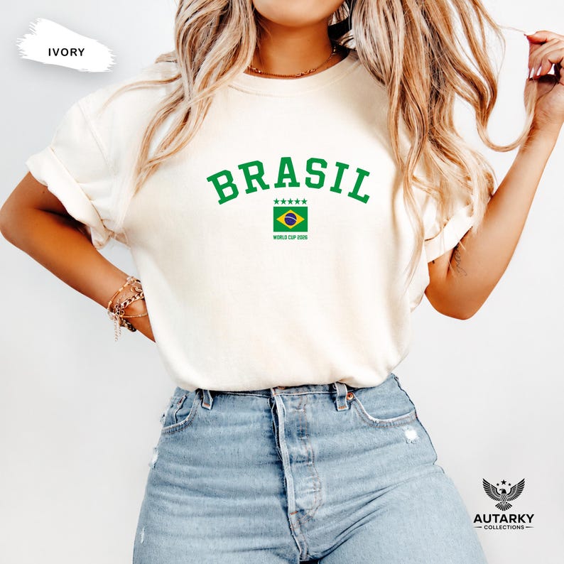 May include: An ivory t-shirt featuring the green text BRASIL in an arched font above a small graphic of the Brazilian flag, five green stars, and the text WORLD CUP 2026. The shirt is paired with light blue denim jeans.