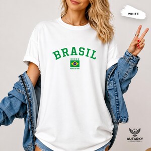 May include: A white short-sleeved t-shirt featuring the green text BRASIL in an arched font above a small Brazilian flag graphic with five green stars and the text WORLD CUP 2026 underneath, worn with a blue denim jacket over the shoulders.