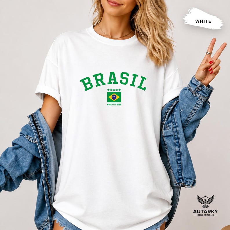 May include: A white short-sleeved t-shirt featuring the green text BRASIL in an arched font above a small Brazilian flag graphic with five green stars and the text WORLD CUP 2026 underneath, worn with a blue denim jacket over the shoulders.