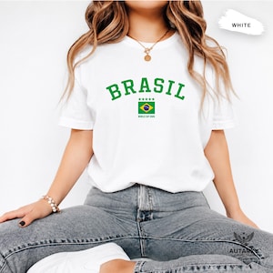 May include: A white short-sleeve t-shirt featuring the green text BRASIL in an arched font above a graphic of the Brazilian flag with five green stars and the text WORLD CUP 2026 printed in small green letters underneath the flag.