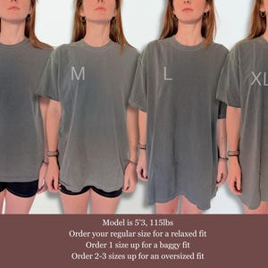 May include: Four charcoal grey short-sleeve t-shirts in sizes S, M, L, and XL. The shirts feature a crew neckline and a relaxed fit. Text on the image includes size labels S, M, L, XL and sizing advice: Model is 5'3, 115lbs. Order your regular size for a relaxed fit. Order 1 size up for a baggy fit. Order 2-3 sizes up for an oversized fit.
