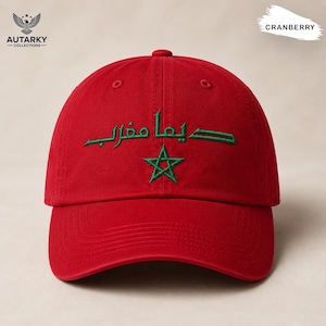 May include: A cranberry red baseball cap featuring green embroidered Arabic text and a green five-pointed star centered on the front panel. The cap has a structured crown, a curved brim, and stitched eyelets for ventilation on the top panels.