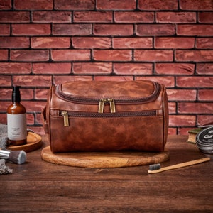 Op de afbeelding: Een bruine leren toilettas voor reizen met twee vakken met rits. De tas ligt op een houten oppervlak met een tandenborstel, een fles lotion en een klein potje cr&egrave;me.