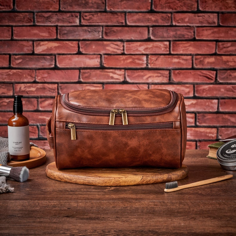Op de afbeelding: Een bruine leren toilettas voor reizen met twee vakken met rits. De tas ligt op een houten oppervlak met een tandenborstel, een fles lotion en een klein potje cr&egrave;me.