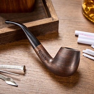 May include: A wooden tobacco pipe with a dark brown finish and a black stem. The name "Noah" is engraved on the pipe's stem. The pipe is resting on a wooden surface, with cleaning tools and a wooden tray in the background.