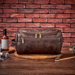 Op de afbeelding: Een bruine leren reistas voor toiletartikelen met twee ritsvakken en gouden ritsen. De tas ligt op een houten oppervlak.