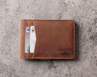 May include: A brown leather wallet with the name "Jacob" and a mustache graphic engraved on the front. The wallet has card slots and is open, revealing credit cards. The wallet is on a gray surface.