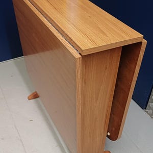 May include: A light brown, wooden drop-leaf table with a rectangular top and a folding leaf. The table has a simple design with four small legs. The table is approximately 76 cm long when folded.