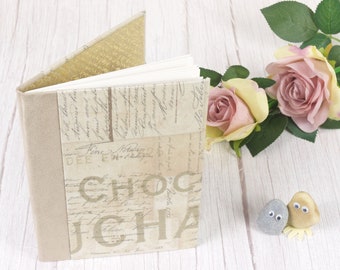 May include: A beige linen-covered journal with a gold-toned spine and a decorative cover featuring a vintage-style print with the word "Chocolat" in large letters. The journal is sitting on a white wooden surface with a bouquet of pink roses in the background.