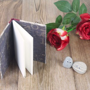 May include: A small, brown, fabric-covered journal with white pages is open on a wooden surface. Two grey stones with painted eyes are in the foreground. Two red roses with green leaves are in the background.