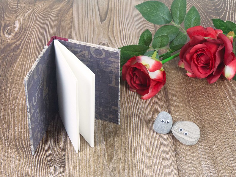 May include: A small, brown, fabric-covered journal with white pages is open on a wooden surface. Two grey stones with painted eyes are in the foreground. Two red roses with green leaves are in the background.