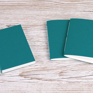 May include: Three teal notebooks with white-edged pages are arranged on a light wood-grain surface. One notebook is positioned separately, while the other two are slightly overlapping. The covers have a textured appearance.