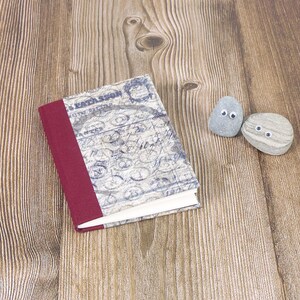 May include: A small, red-bound notebook with a vintage-style cover featuring blue and white script. Two small grey stones with googly eyes are placed on a wooden surface.