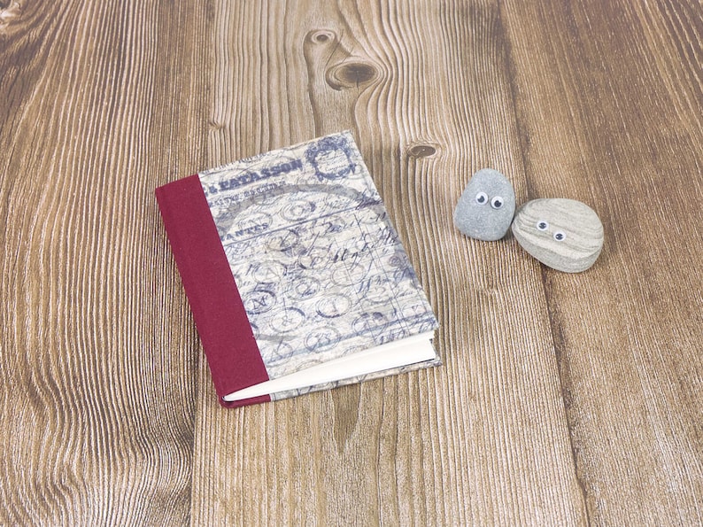 May include: A small, red-bound notebook with a vintage-style cover featuring blue and white script. Two small grey stones with googly eyes are placed on a wooden surface.