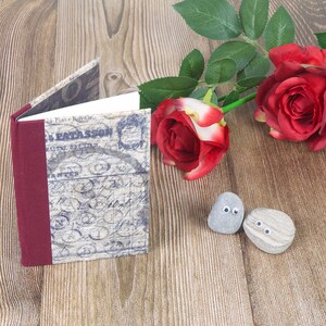 May include: A small, vintage-style journal with a burgundy spine and a blue and white patterned cover. The journal is sitting on a wooden surface with two grey stones with googly eyes and a red rose with green leaves.