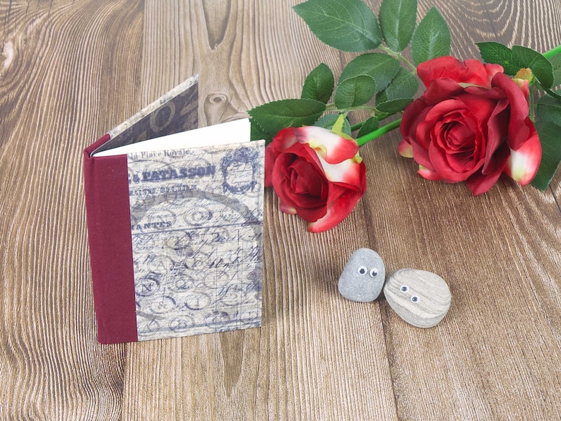 May include: A small, vintage-style journal with a burgundy spine and a blue and white patterned cover. The journal is sitting on a wooden surface with two grey stones with googly eyes and a red rose with green leaves.