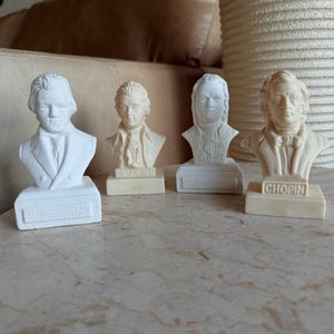 May include: Four small, off-white busts of famous composers: Beethoven, Mozart, and Chopin. Each bust is mounted on a rectangular base with the composer's name and birth year. The busts are arranged on a marble surface.