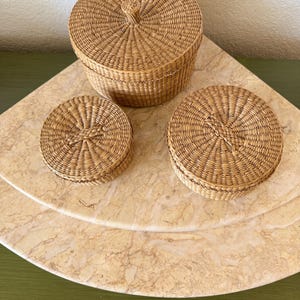 May include: Three round, woven wicker baskets with matching lids. One basket is taller and larger than the other two, which are smaller and flatter. Each basket features a light tan, natural fiber construction with a tight, circular weave pattern.