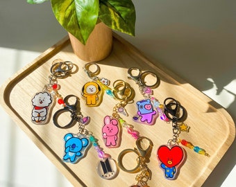 May include: A collection of colorful acrylic keychains featuring cartoon characters with beaded accents. The keychains are displayed on a wooden tray with a small potted plant in the background. Each keychain has a different character design.